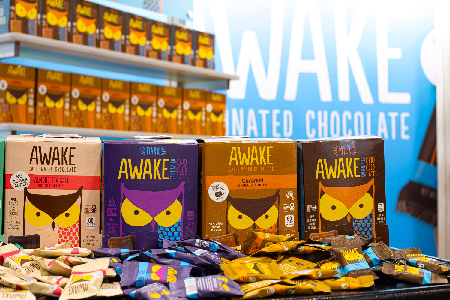 Boxes of Awake caffeinated chocolate with owl illustrations stand on a table of wrapped chocolates in front of a large Awake sign