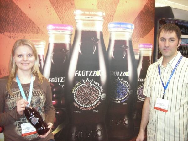 A woman holding a dark Frutzzo pomegranate juice bottle and a man in a striped shirt stand in front of a large backdrop of Frutzzo bottles. Both wear lanyards and badges.