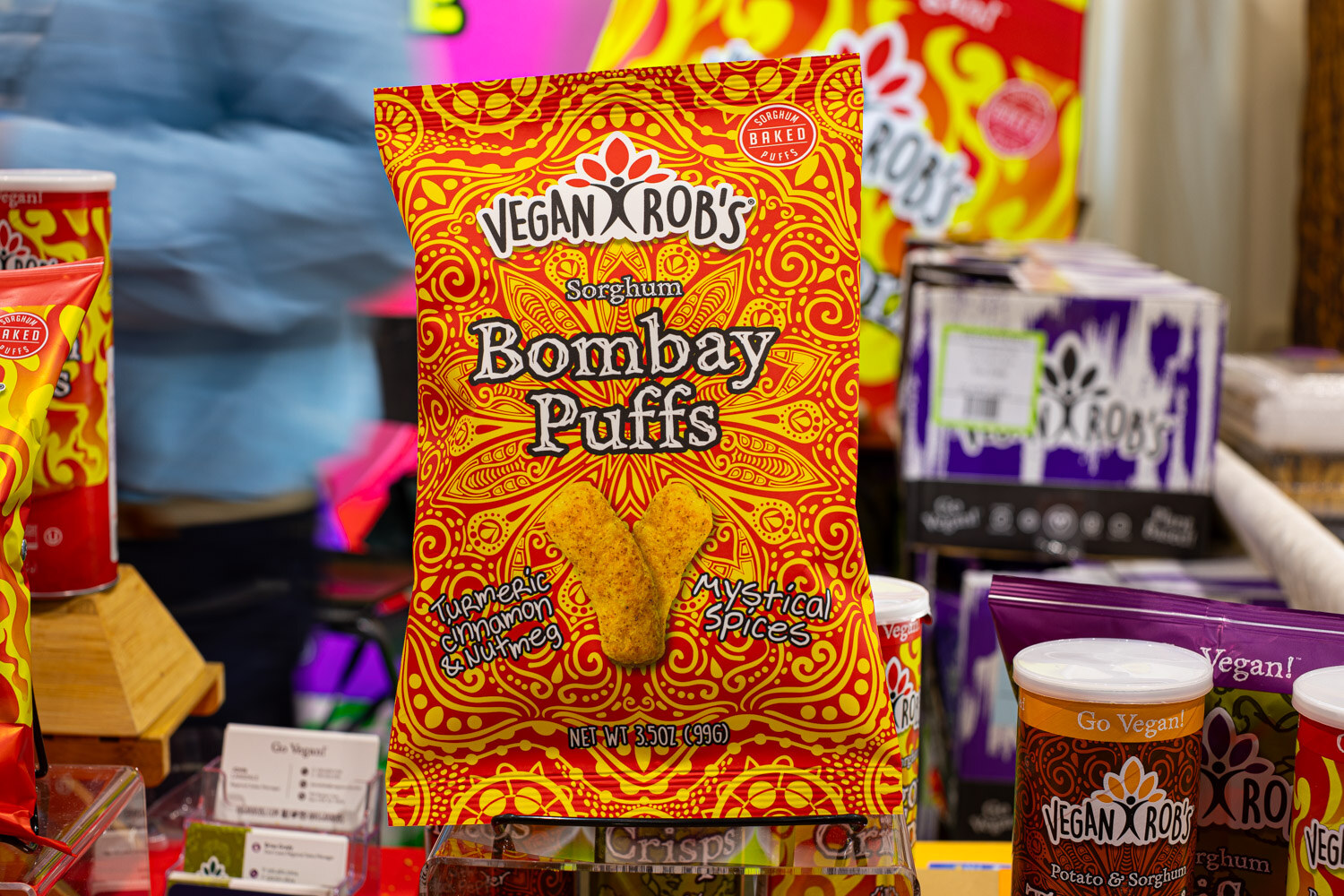A Vegan Rob's Sorghum Bombay Puffs bag in red and gold stands among other Vegan Rob's products
