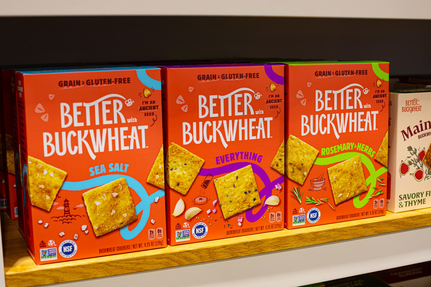 Orange boxes of Better Buckwheat crackers in sea salt, everything and rosemary herb flavors on a wooden shelf