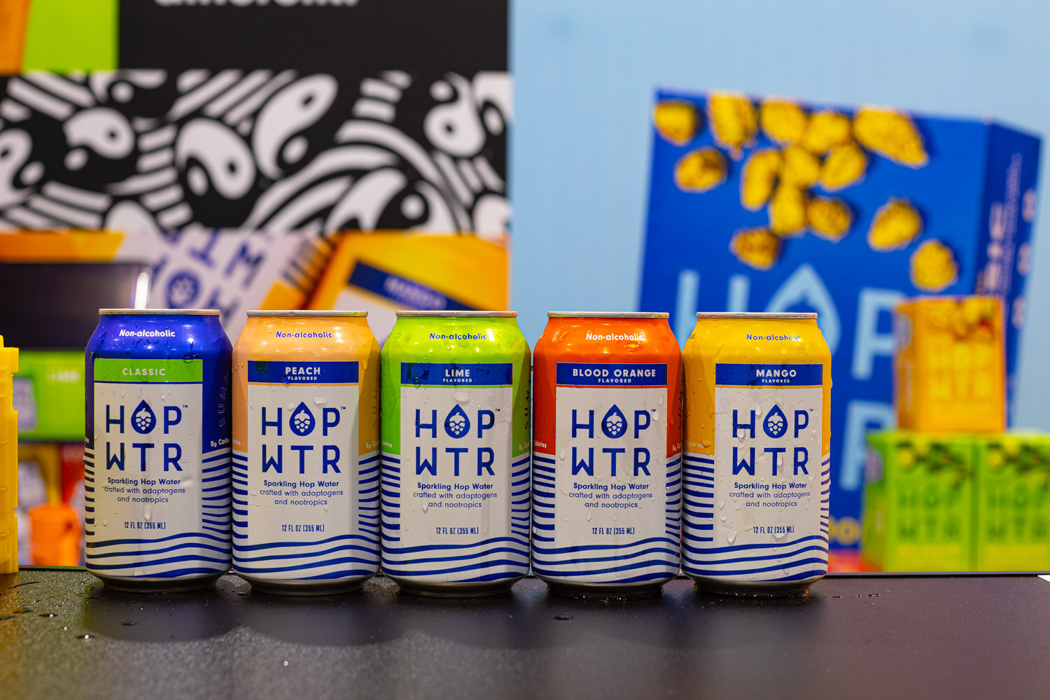 Five HOP WTR sparkling hop water cans with white and blue labels line up in front of a bold black and white pattern