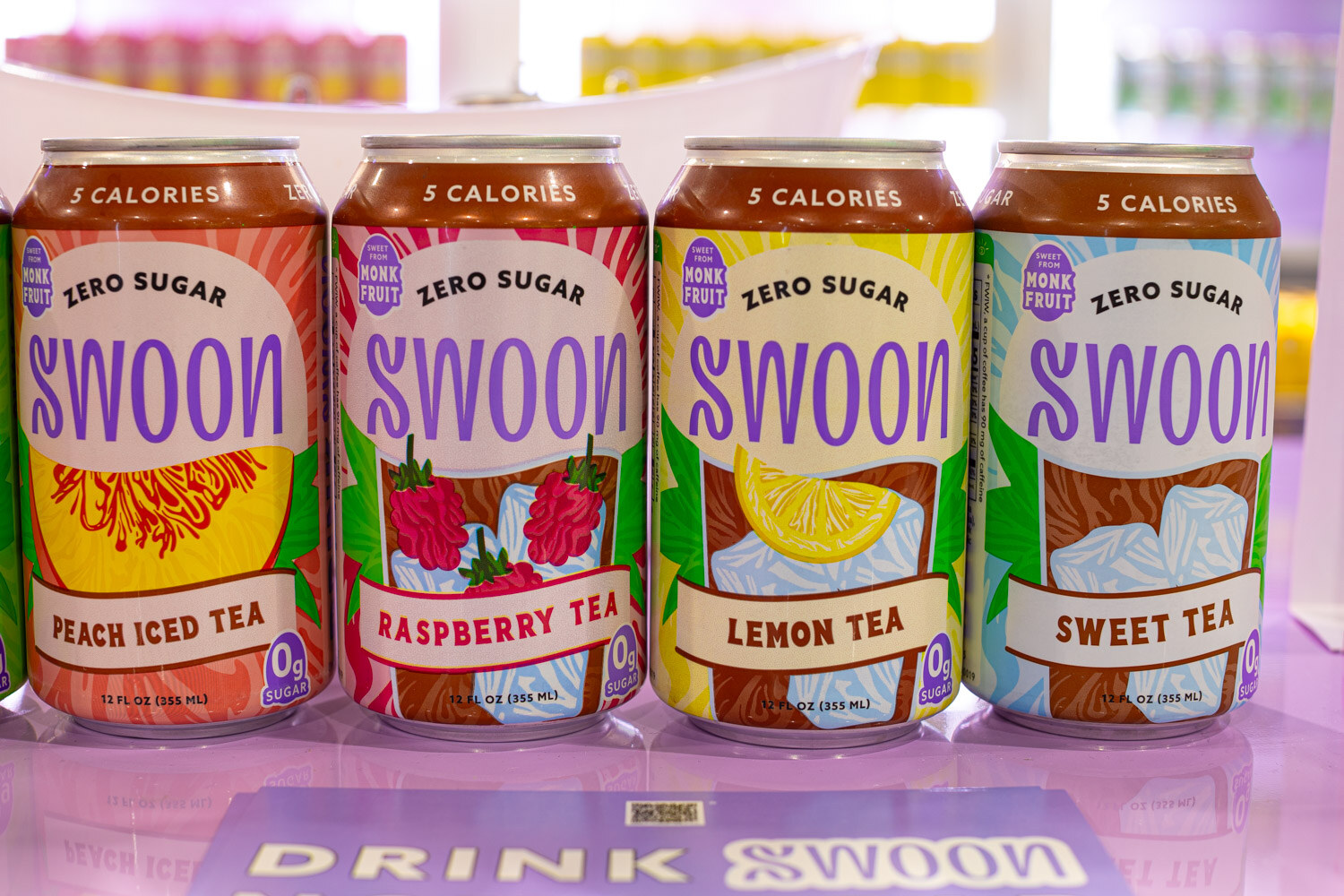 Four Swoon zero sugar tea cans in peach, raspberry, lemon and sweet tea flavors on a purple-lit display
