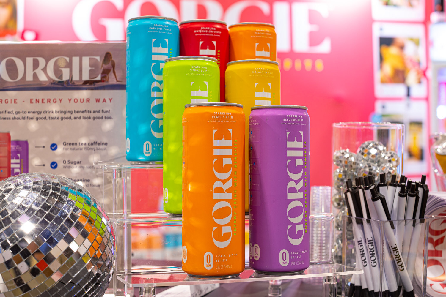 Stacked Gorgie energy cans in teal, orange, yellow and purple stand beside a mirrored disco ball and a stand of pens