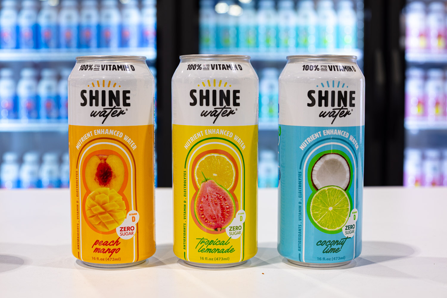 Three Shine Water cans in peach mango, tropical lemonade and coconut lime flavors stand in front of a cooler
