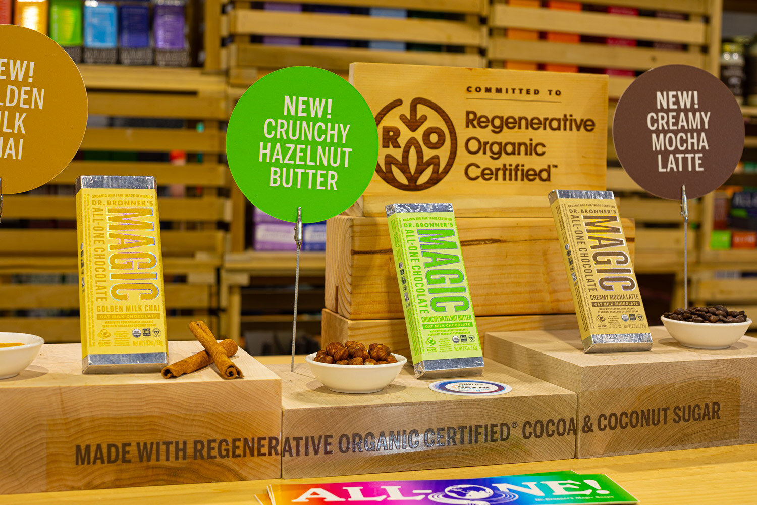 Gold and green MAGIC all-one chocolate bars stand on a wooden display with New Crunchy Hazelnut Butter and Regenerative Organic Certified signs