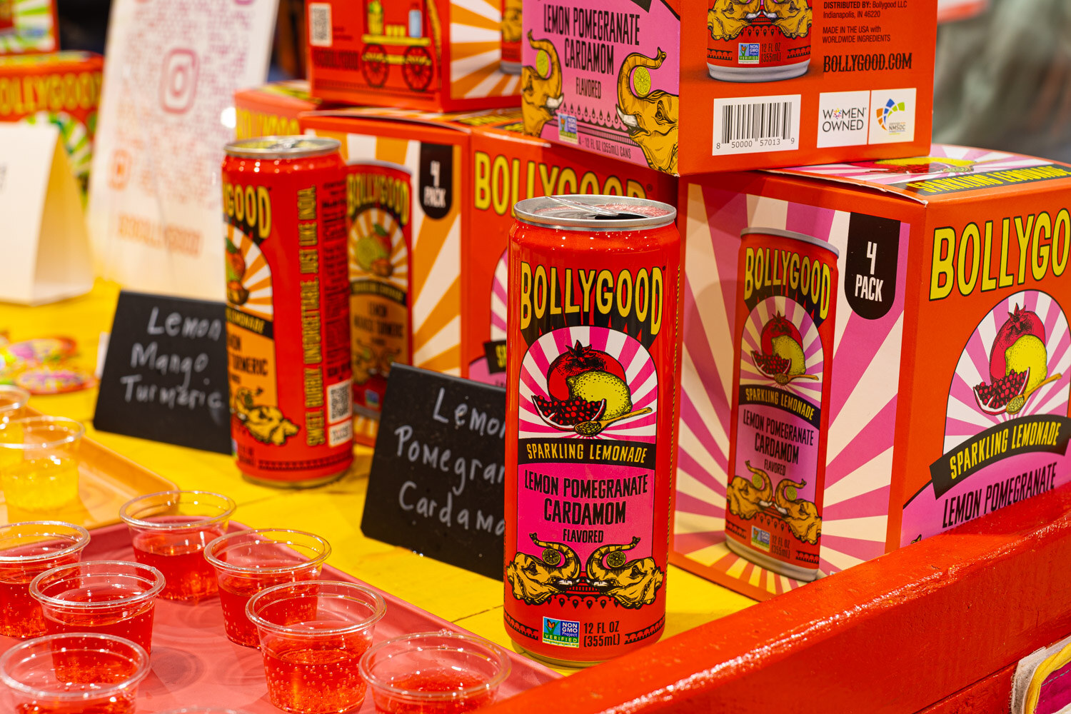 Bollygood sparkling lemonade cans in colorful packaging stand above small sample cups and a sign for lemon mango turmeric