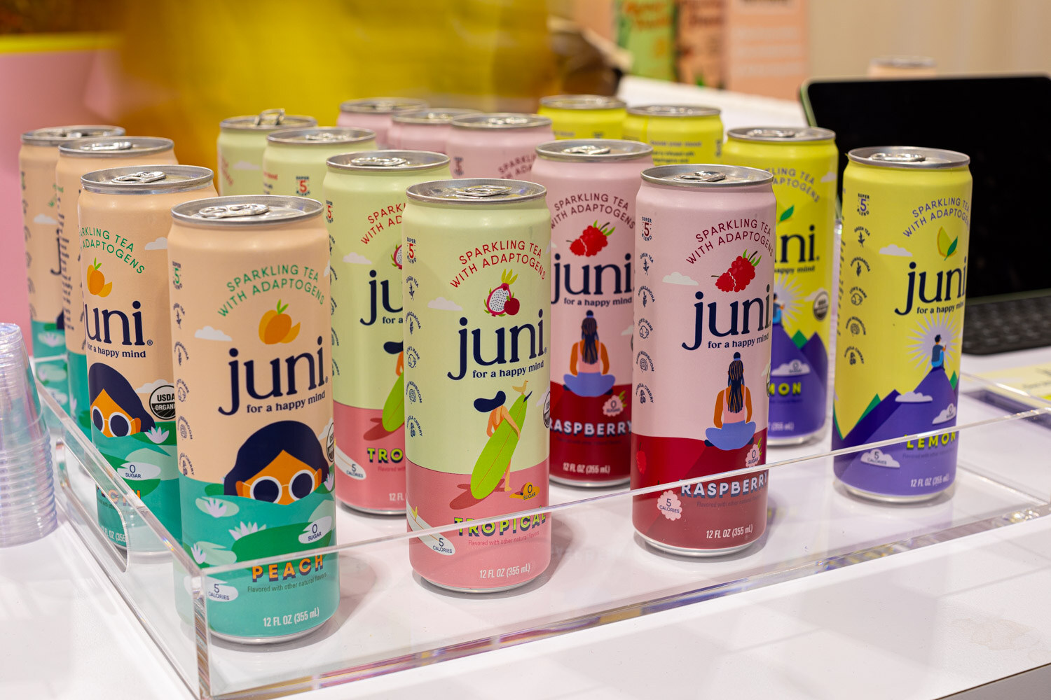 Colorful Juni sparkling tea cans with illustrated characters sit on a clear acrylic stand