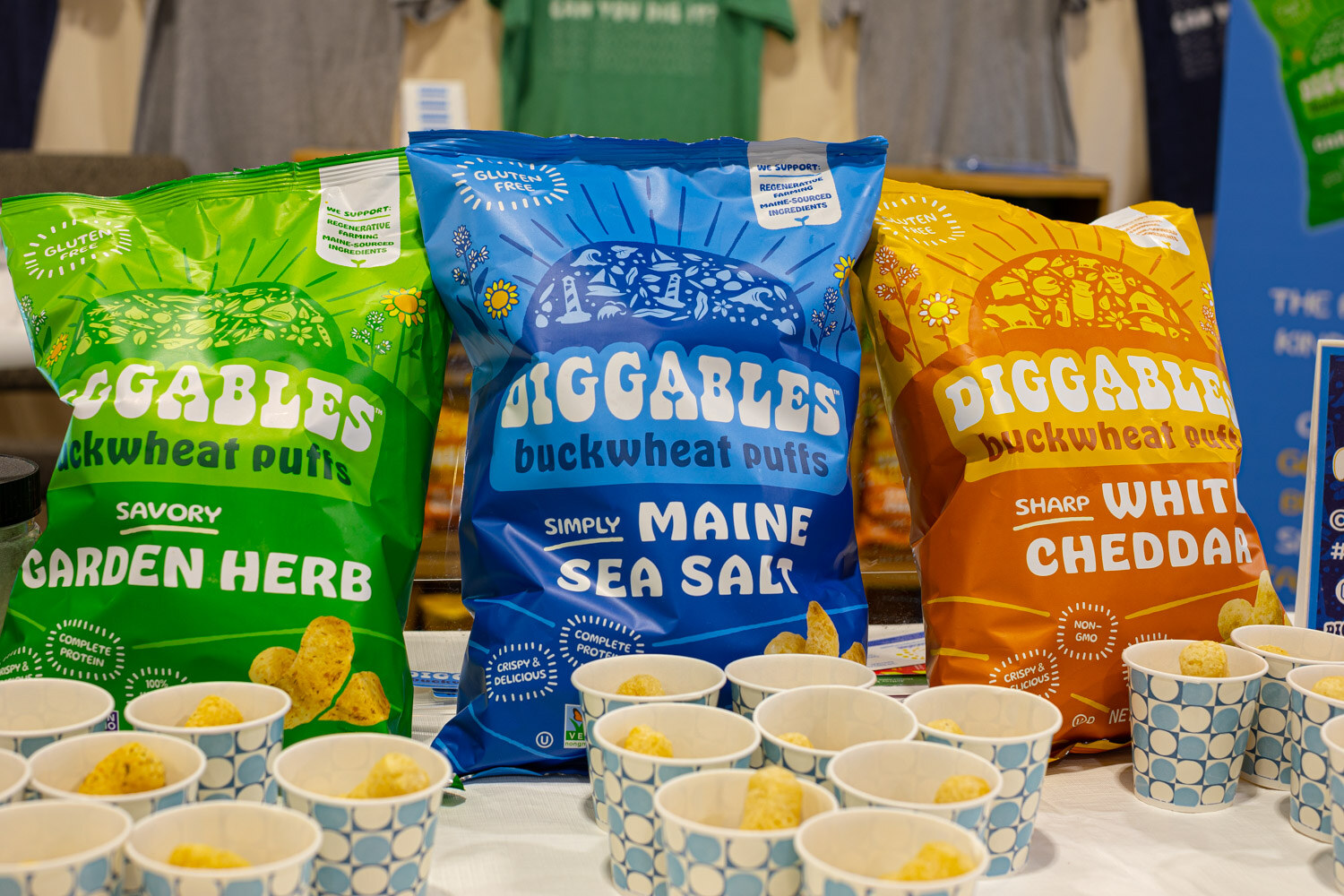 Three Diggables buckwheat puffs bags in Garden Herb, Maine Sea Salt and White Cheddar flavors stand above sample cups