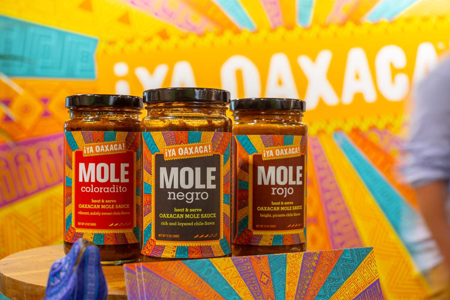 Jars of Mole Coloradito, Mole Negro and Mole Rojo stand in front of a bright Ya Oaxaca banner