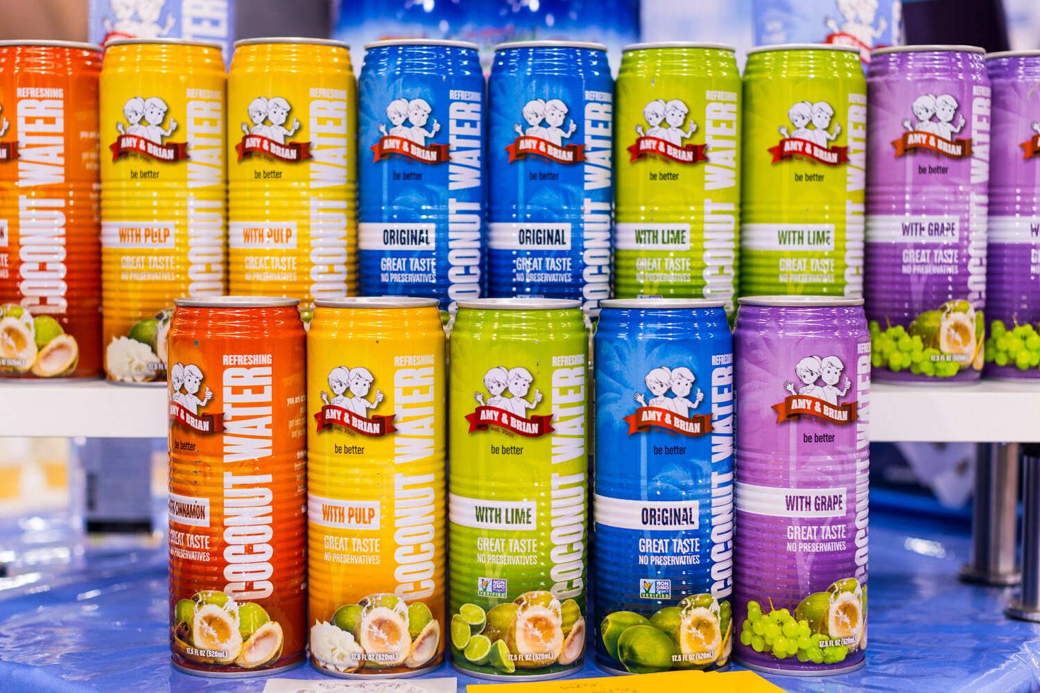 Two rows of orange, yellow, blue, green and purple Amy & Brian coconut water cans in several flavors.