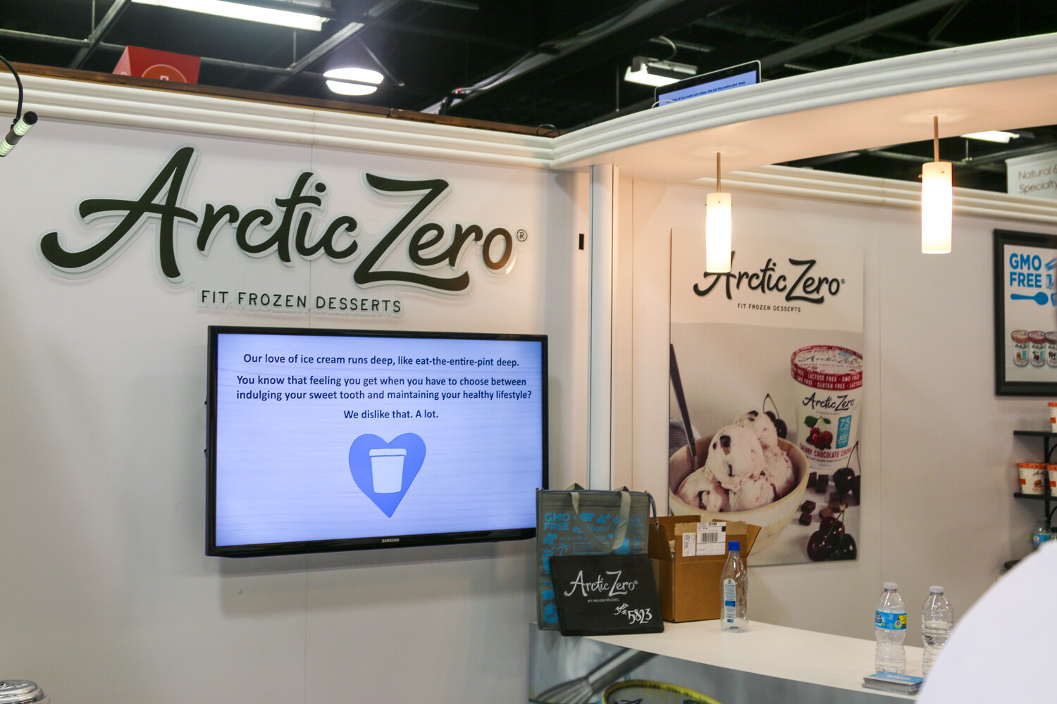 White booth wall with an Arctic Zero Fit Frozen Desserts sign, a monitor with text about ice cream and a pint poster.
