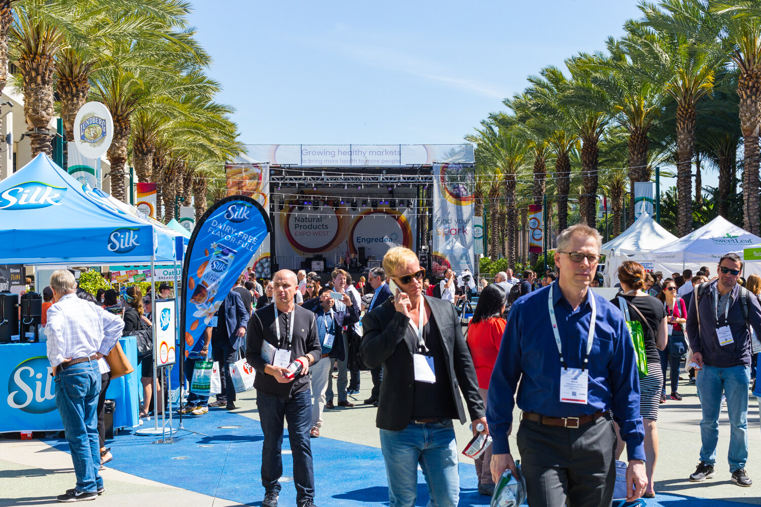 Attendees with badges walking in a sunny plaza with a blue Silk tent at left and a stage with Natural Products Expo West signage behind them.