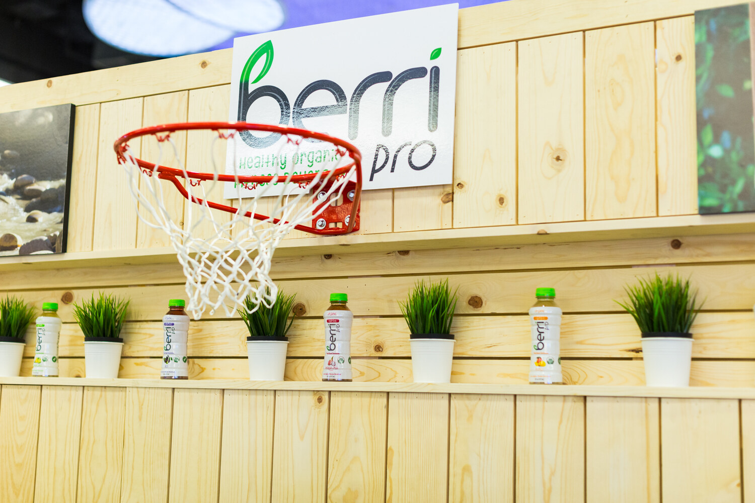Wood-plank booth wall with a Berri Pro sign and a basketball hoop above a shelf of green-capped bottles and small potted grass.