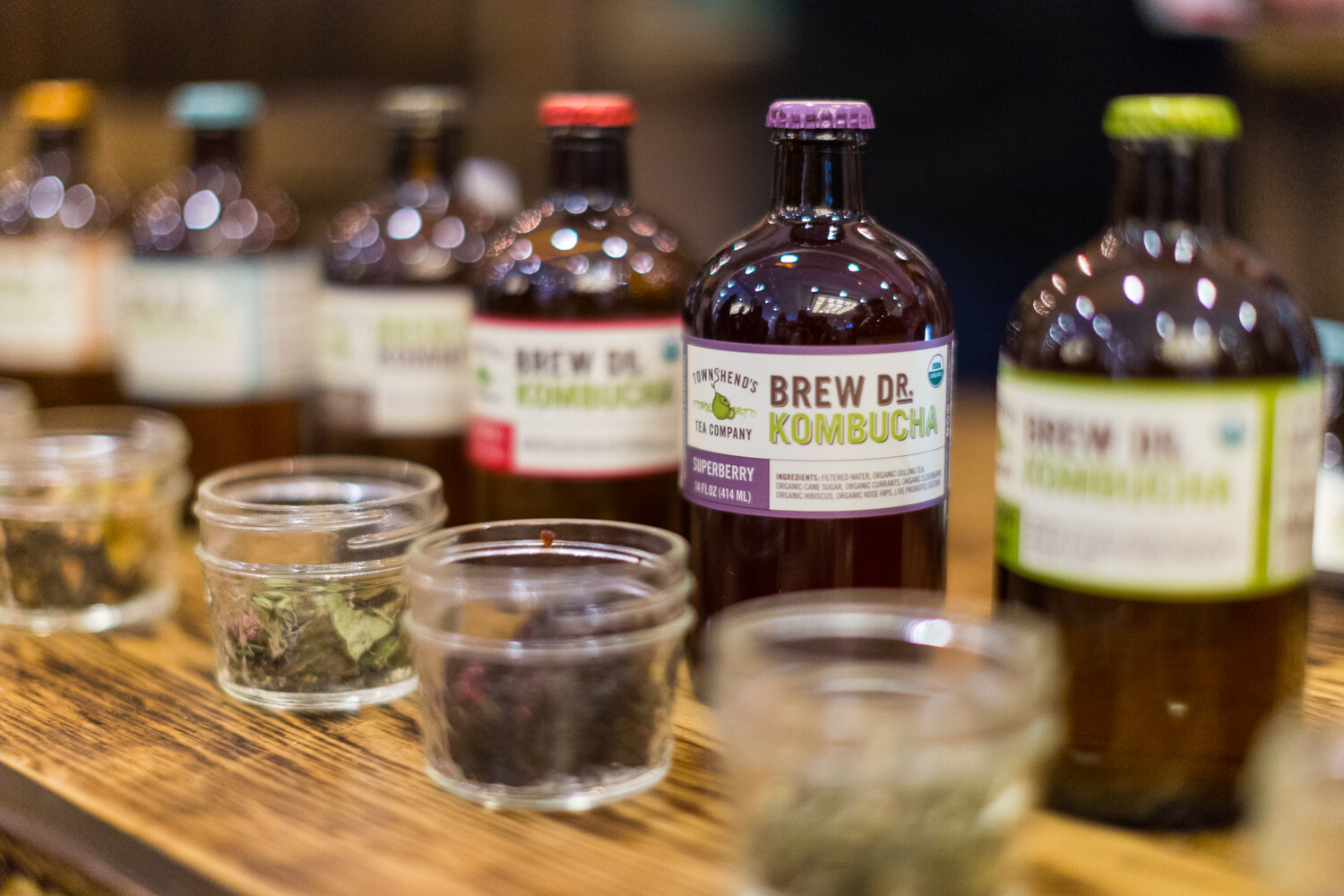 Row of Brew Dr. Kombucha bottles with a Superberry label in focus and small jars of dried tea in front.
