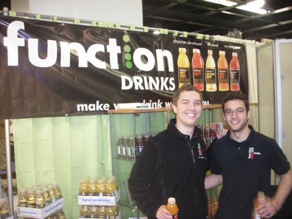 Two smiling men stand at a booth under a black Function Drinks banner, one holding an orange bottle. Cases of bottled drinks sit on shelves and a display behind them.