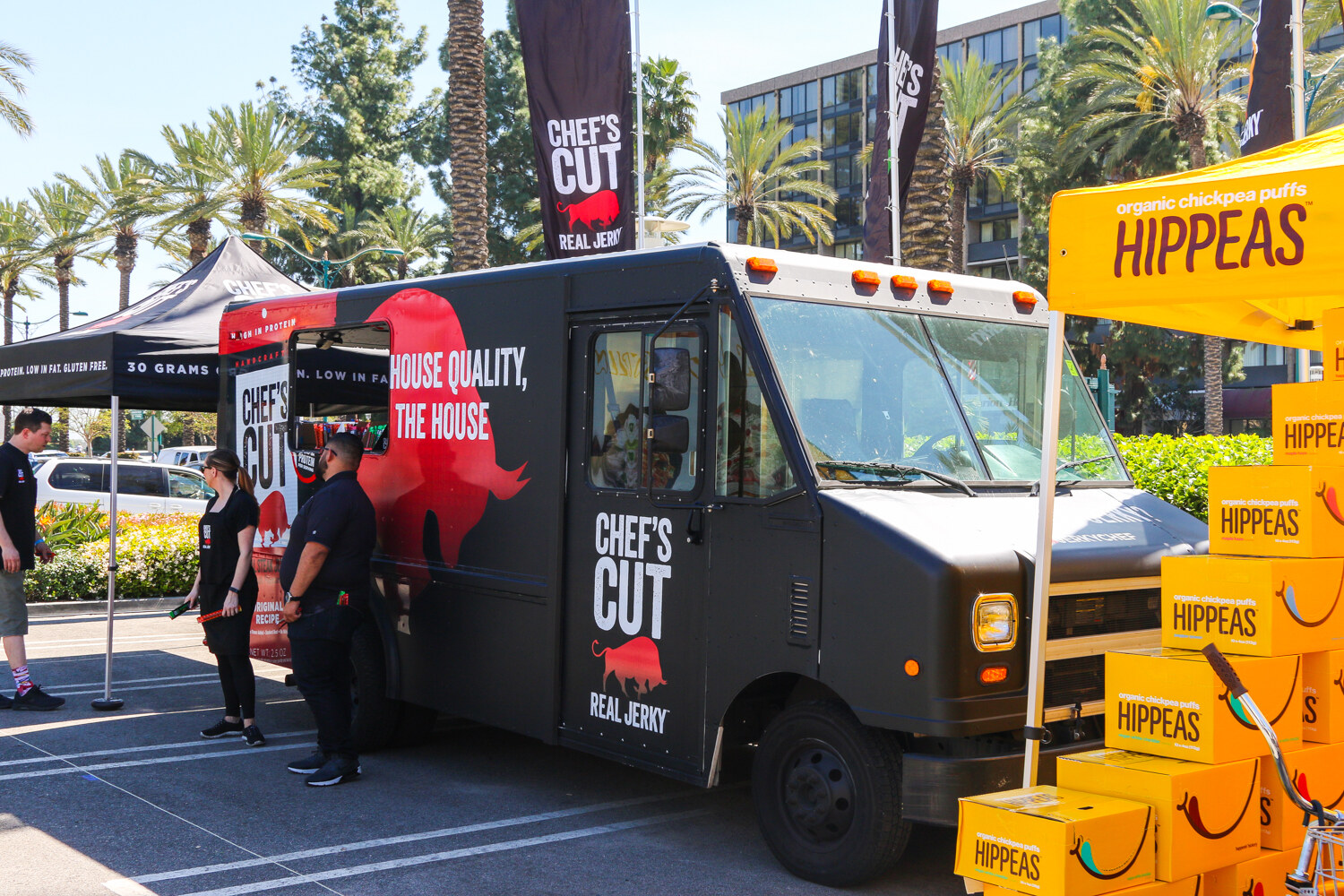 Black Chef's Cut Real Jerky truck with staff standing nearby, and a yellow Hippeas tent with stacked boxes at the right.