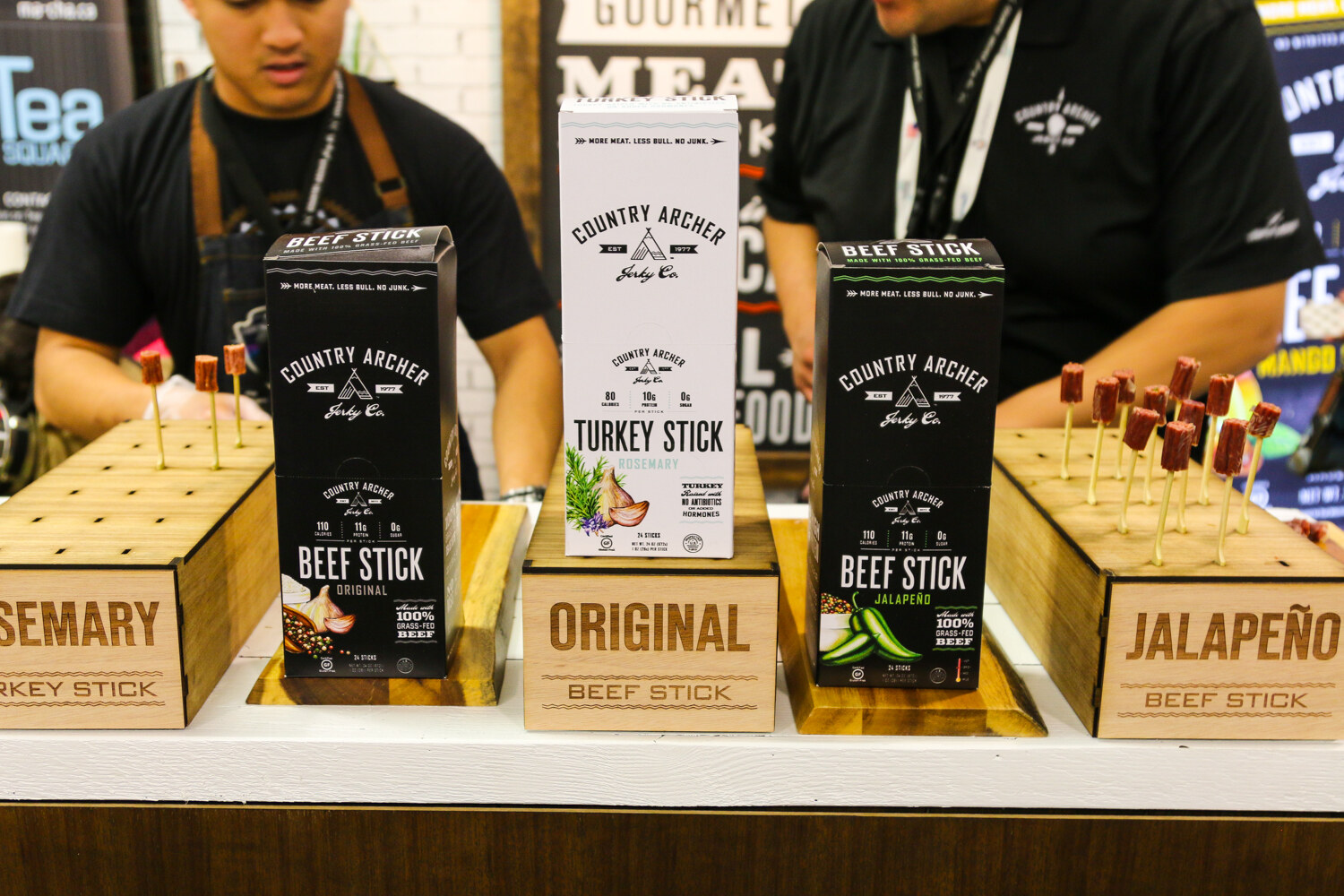 Boxes of Country Archer Original and Jalapeno Beef Stick and Rosemary Turkey Stick on wooden stands with sample skewers, and two staff behind.