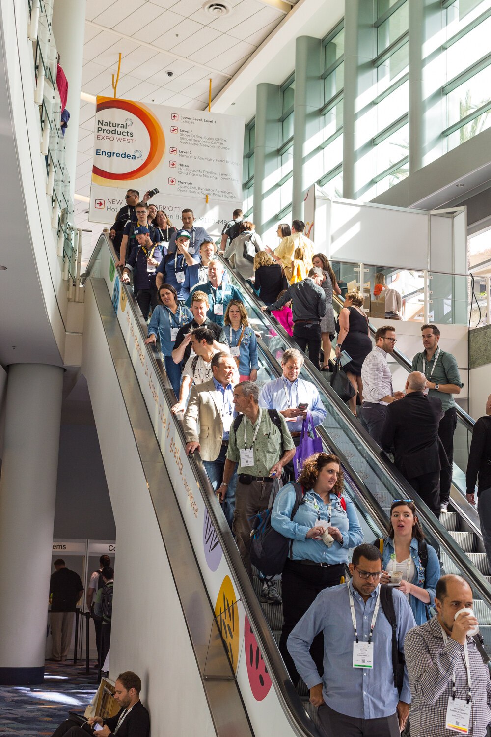 Vertical view of a crowded escalator with badge-wearing attendees and a hanging Natural Products Expo West wayfinding sign.