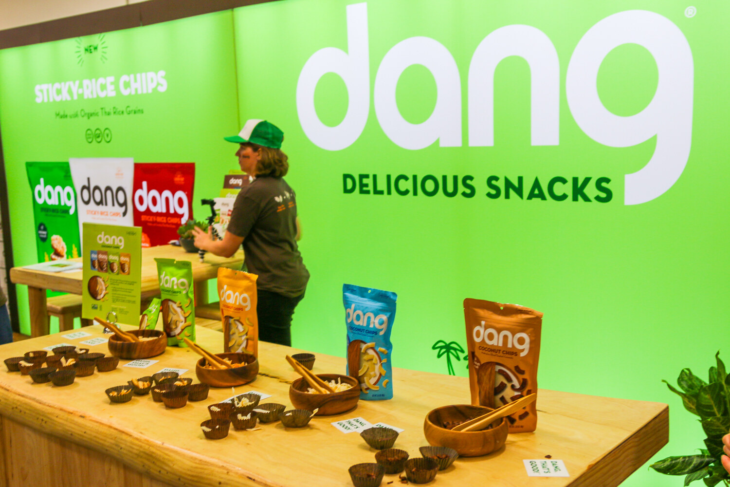 Green booth with a large Dang Delicious Snacks logo, bags of coconut chips and wooden bowls of samples on a long wood counter.