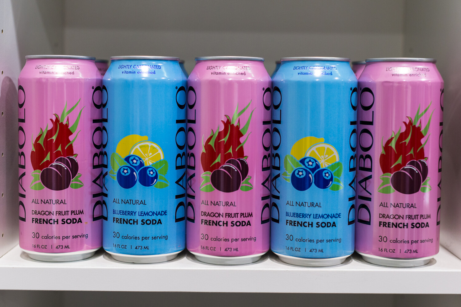 Five Diablo French Soda cans alternating pink dragon fruit plum and blue blueberry lemonade on a white shelf.