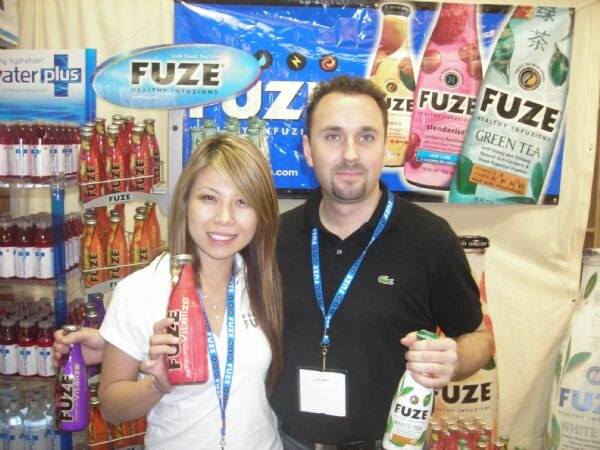 A woman holding a red Fuze bottle and a man in a black Lacoste polo holding a Fuze white tea bottle stand at a booth with Fuze banners and a Water Plus display.