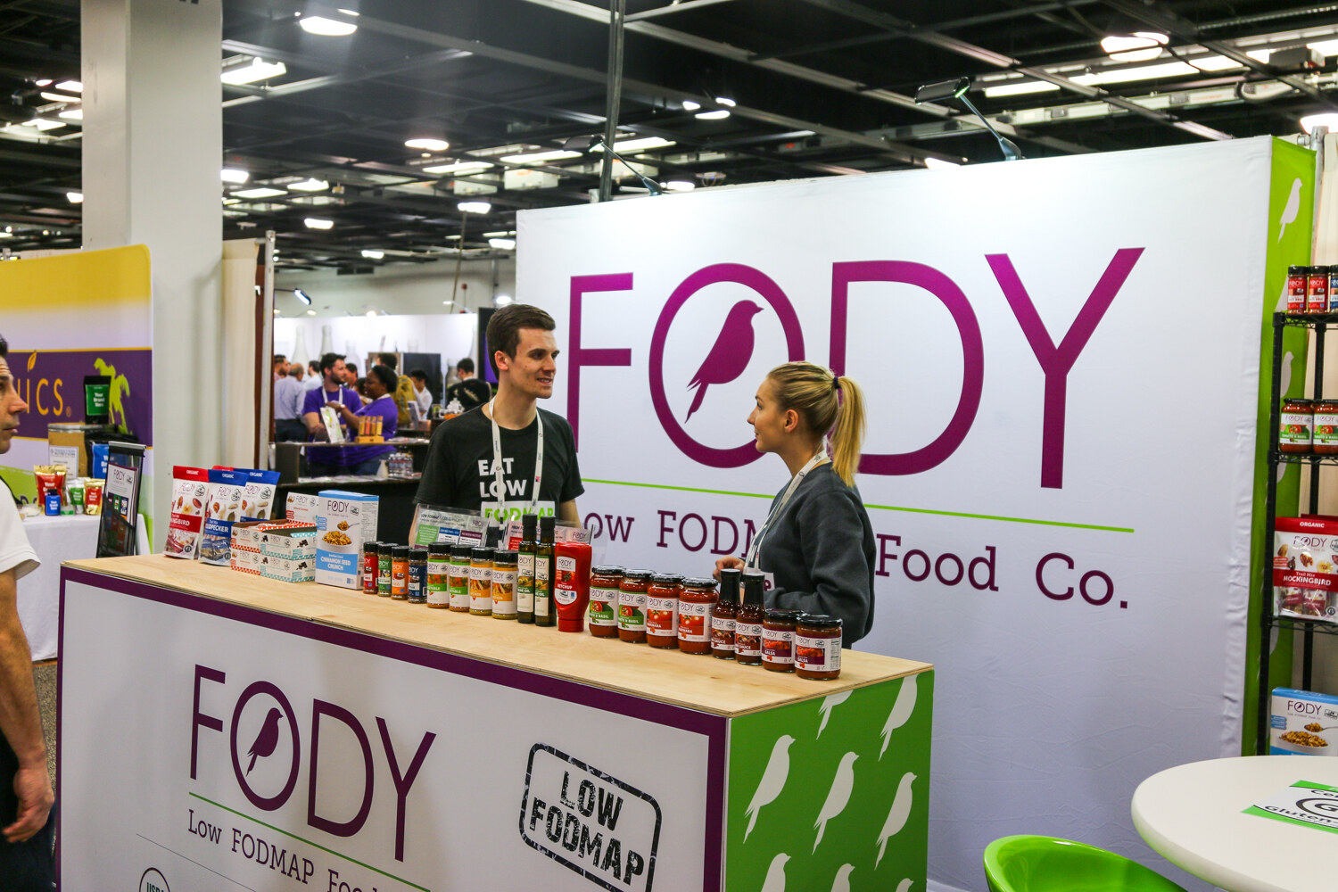 Booth with a large purple Fody Low FODMAP Food Co. wall, a counter of jarred sauces and two staff talking.