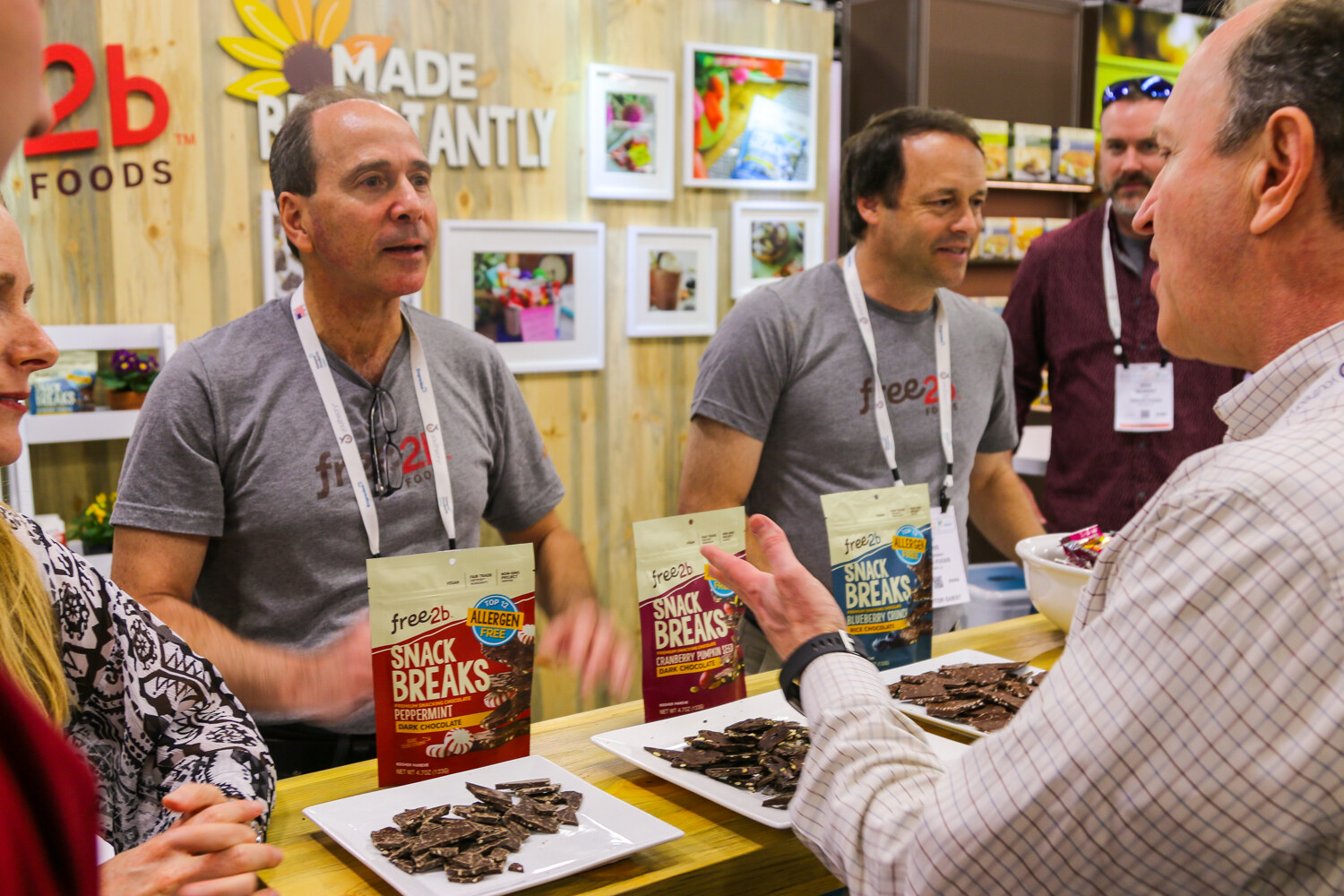 Men in gray free2b T-shirts and lanyards speaking with attendees across a counter with Snack Breaks bags and plates of chocolate bark.