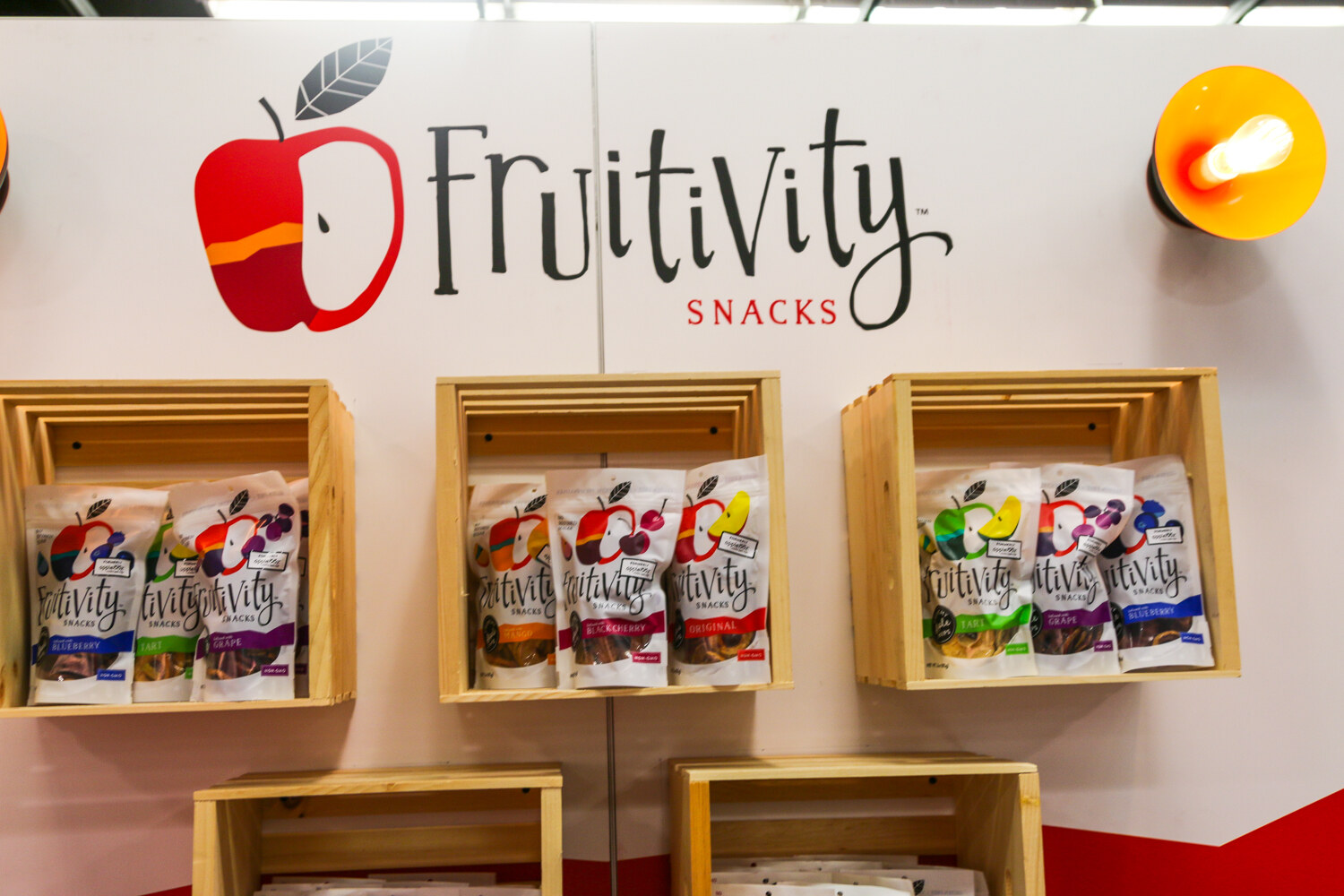 White booth wall with the Fruitivity Snacks apple logo above wooden crates holding bags of dried fruit snacks.