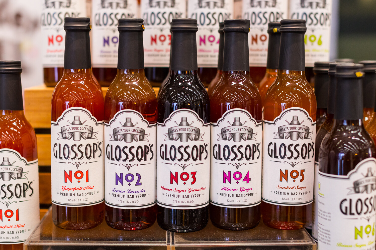Rows of Glossop's Premium Bar Syrup bottles labeled No 1 through No 5 with flavors including Grapefruit Mint and Smoked Sugar.