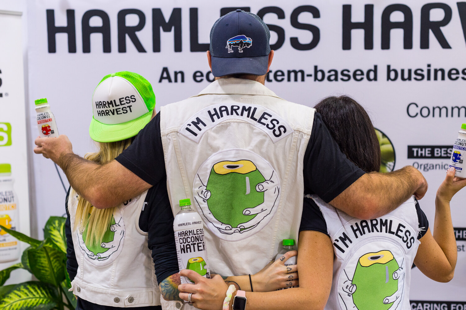 Three people with arms around each other showing I'm Harmless vests, holding bottles of Harmless Harvest coconut water in front of a banner.