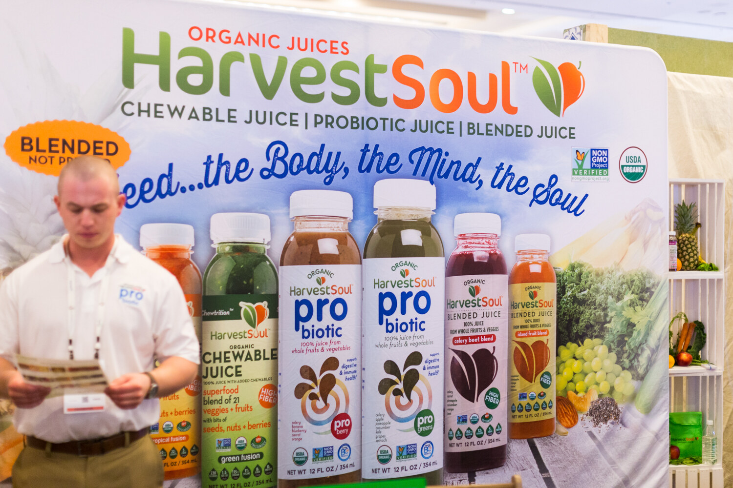 Man in a white polo reading a brochure in front of a banner showing Harvest Soul chewable, probiotic and blended juices.