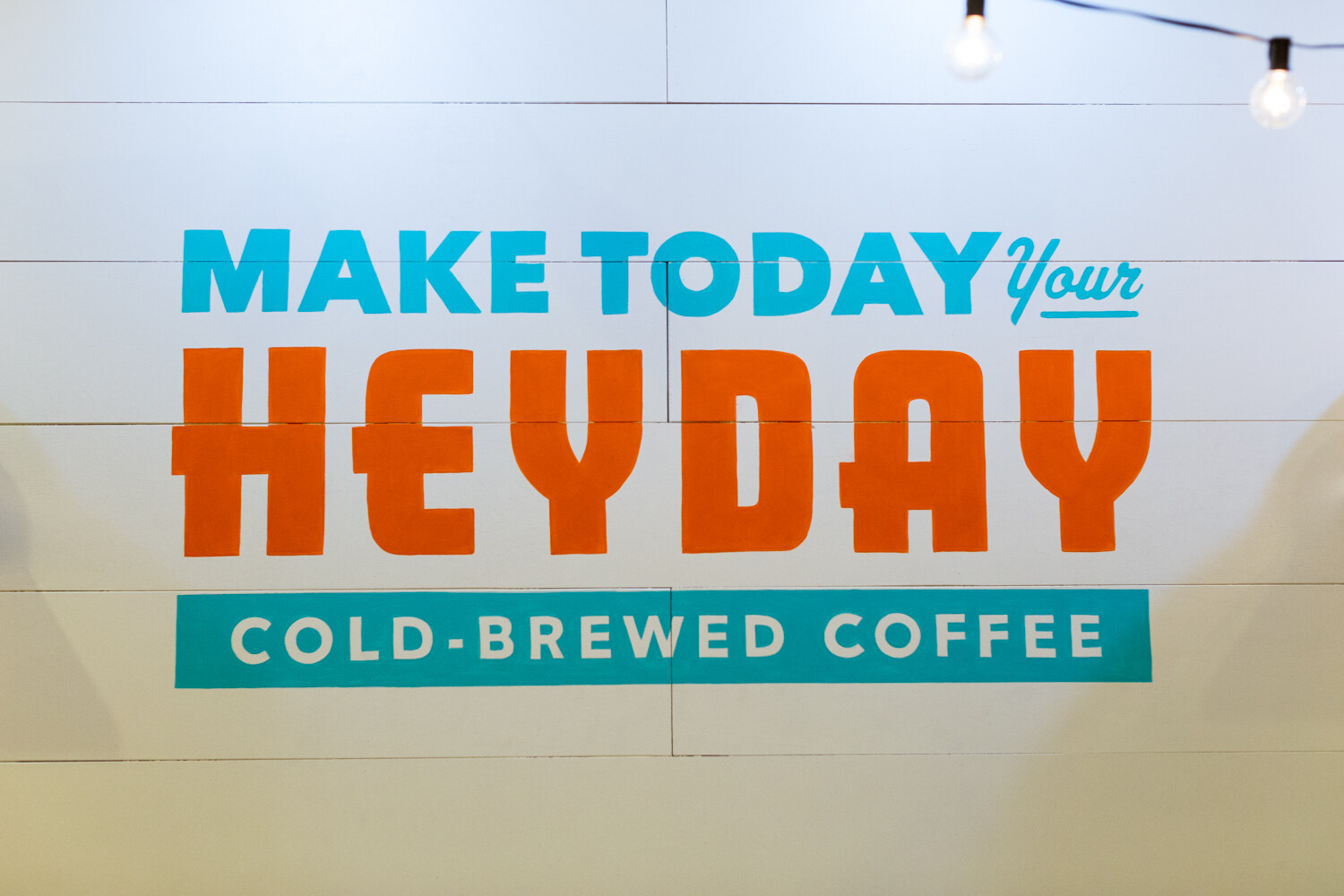 White shiplap wall painted with the words Make Today Your Heyday Cold-Brewed Coffee under string lights.