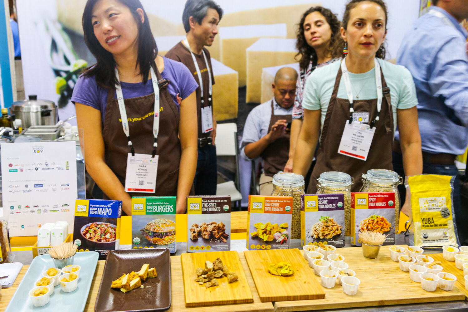 Staff in brown Hodo Soy aprons and badges behind a table of boxed tofu products, cutting boards and small tasting cups.