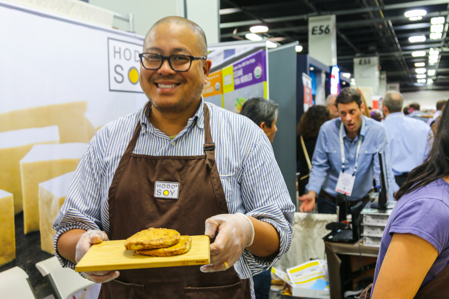 Smiling man with glasses in a striped shirt and brown Hodo Soy apron holding a bamboo board with two tofu patties, show floor behind him.