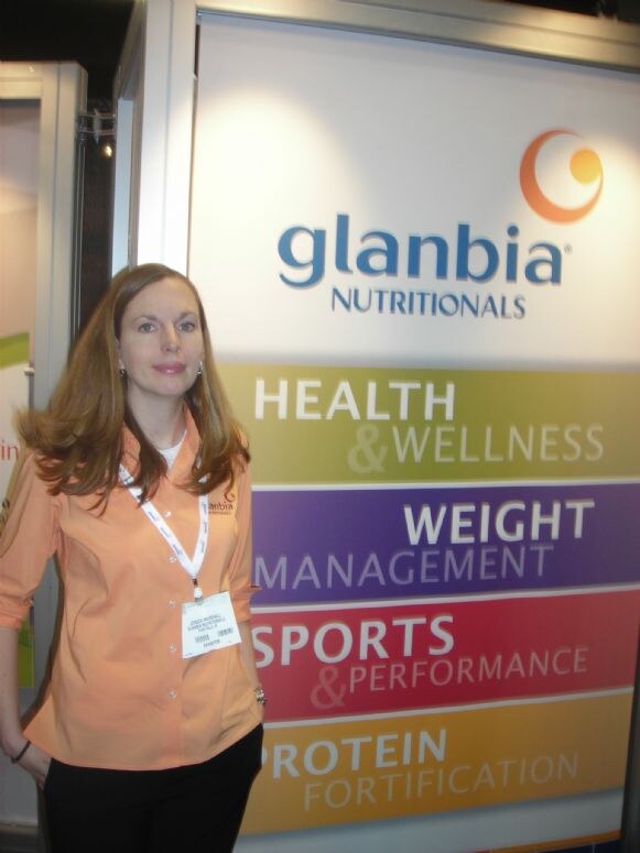 A woman in an orange shirt with a lanyard stands beside a Glanbia Nutritionals sign listing health and wellness, weight management, sports and performance, and protein fortification.