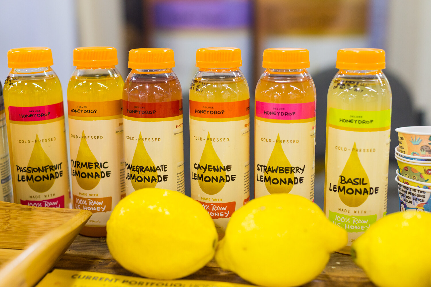 Bottles of Honeydrop lemonade in passionfruit, turmeric, pomegranate, cayenne, strawberry and basil flavors above three whole lemons.