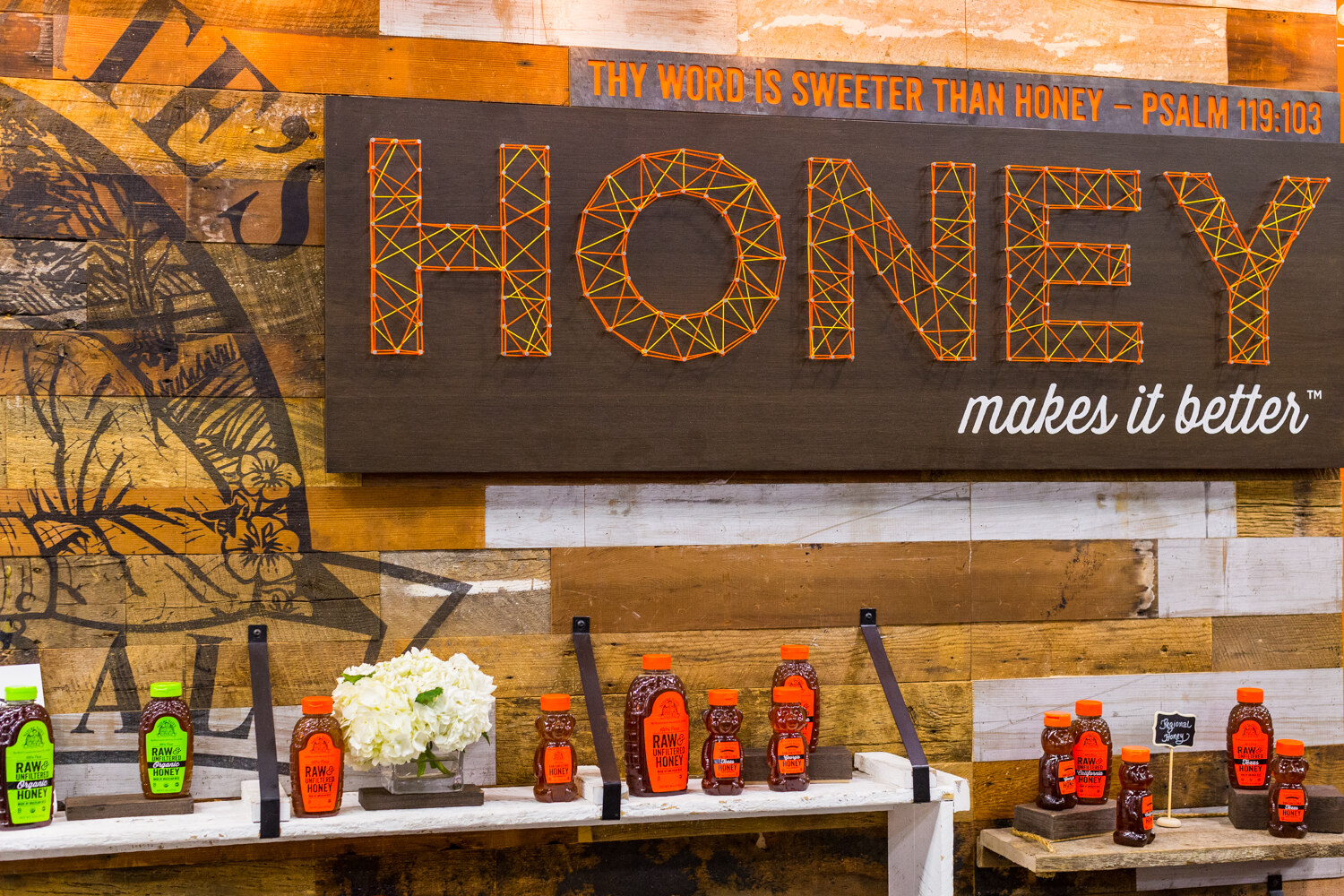 Wooden wall with a large string-art HONEY sign and the words makes it better above bear-shaped honey bottles on shelves.
