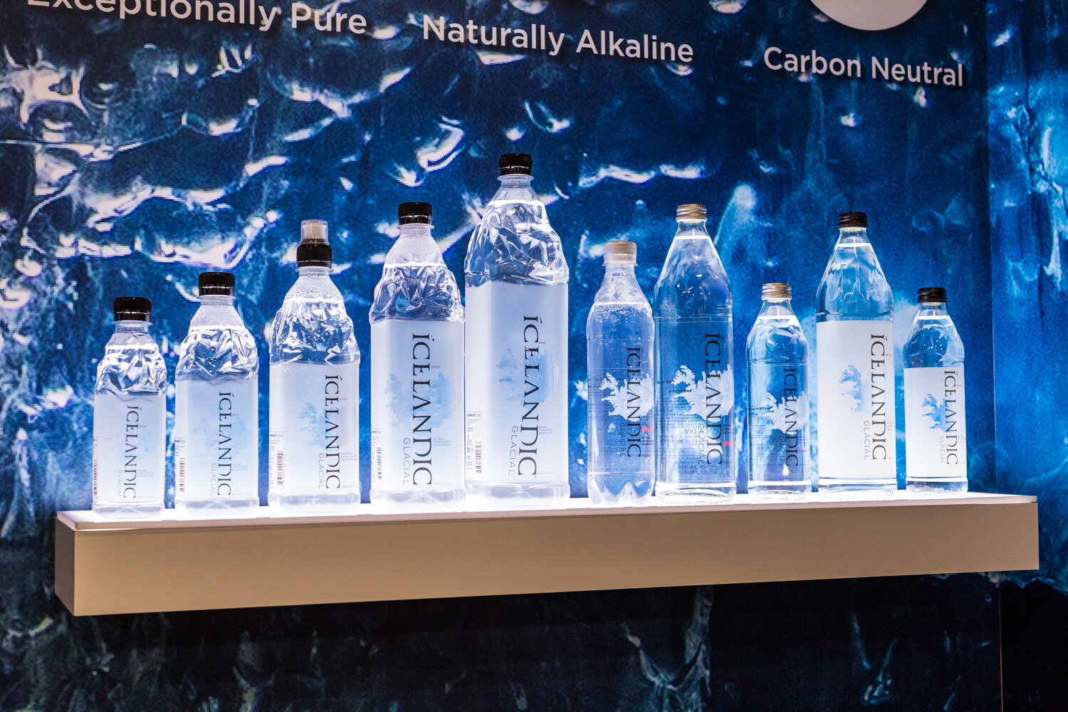 Ten illuminated Icelandic Glacial bottles of different sizes on a shelf in front of a deep blue water graphic.
