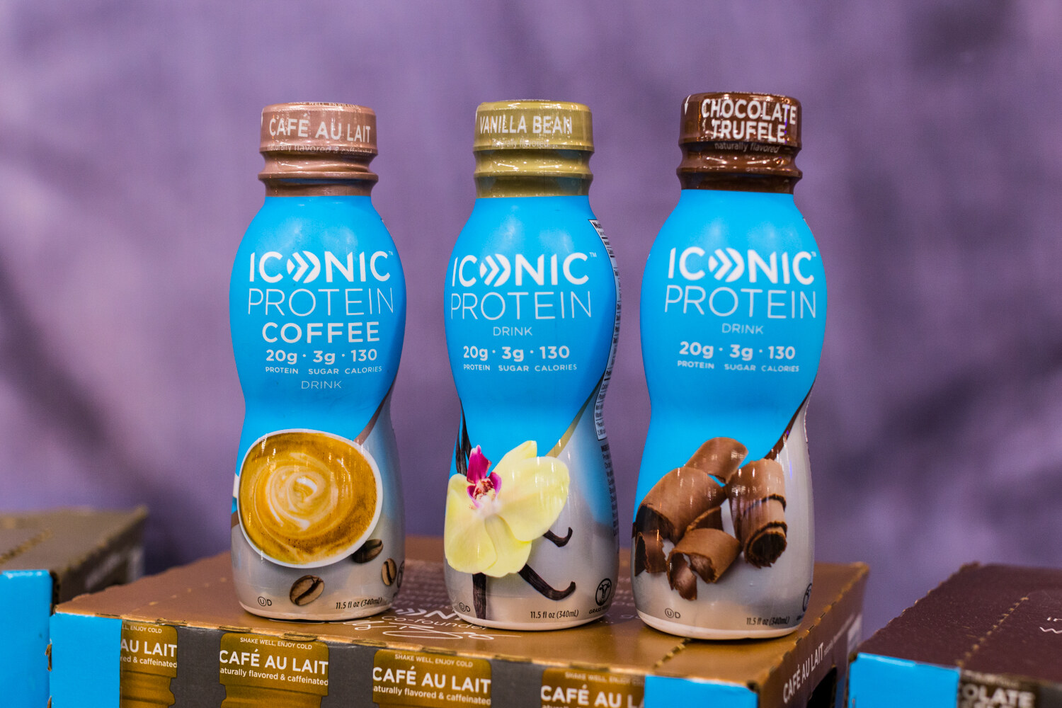 Three blue Iconic Protein bottles in coffee, vanilla bean and chocolate truffle flavors on stacked cases.