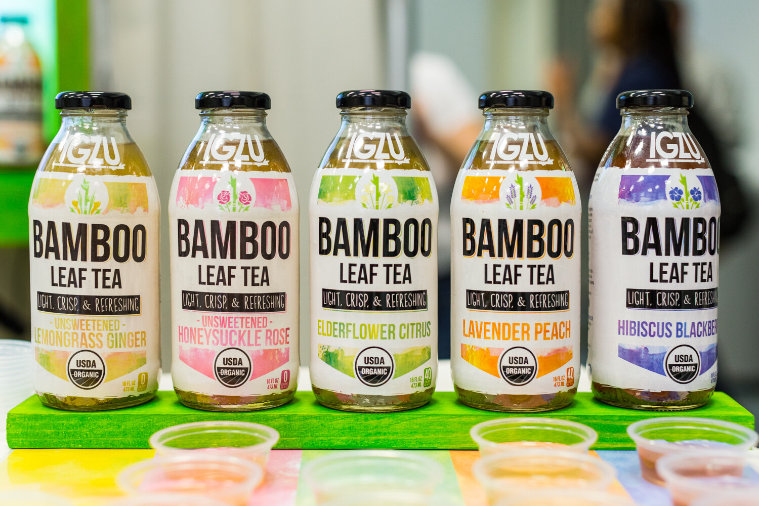 Five bottles of Iglu Bamboo Leaf Tea in flavors such as lemongrass ginger and hibiscus blackberry on a green shelf with sample cups.