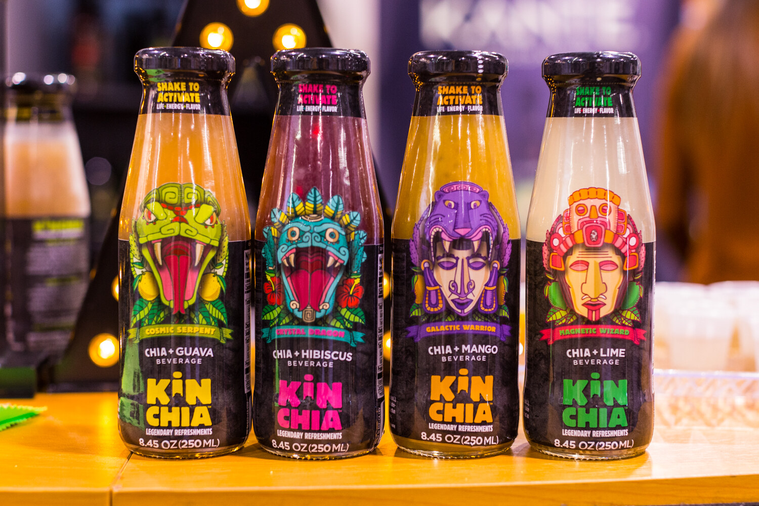 Four glass bottles of Kin Chia chia beverages with guava, hibiscus, mango and lime labels showing masks and a snake.