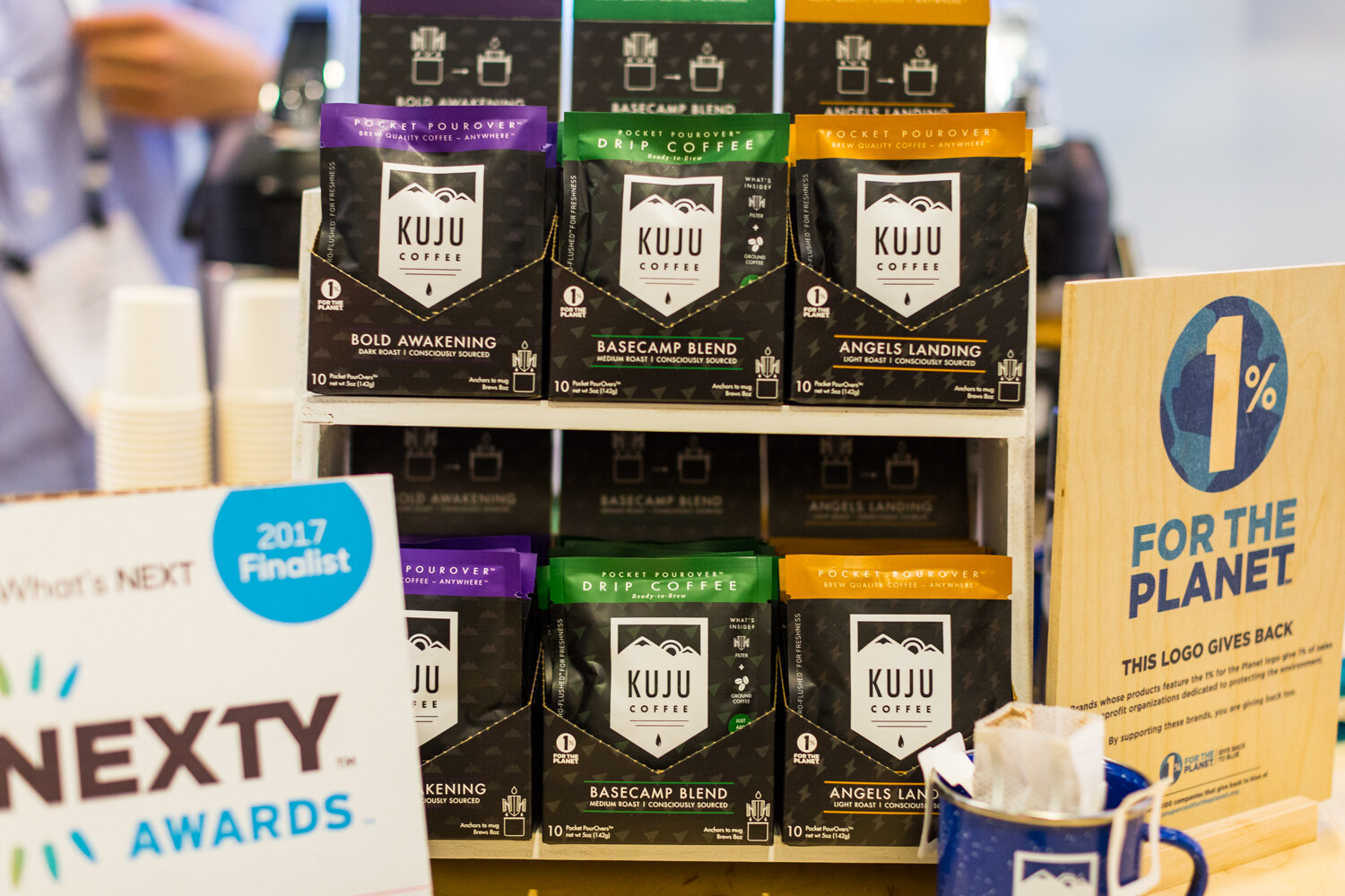 Shelf of Kuju Coffee pourover packs with a 2017 Finalist NEXTY Awards card at left and a 1% For The Planet sign at right.