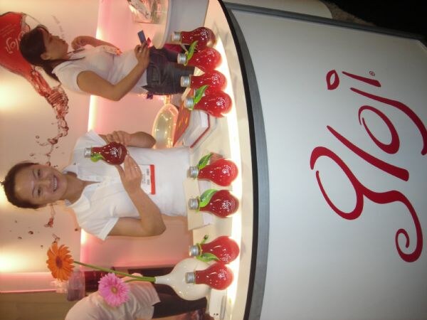 Sideways photo of a Gloji booth where a smiling woman holds a red bulb-shaped bottle and another woman behind her looks at a phone. Similar bottles line a lit curved counter.