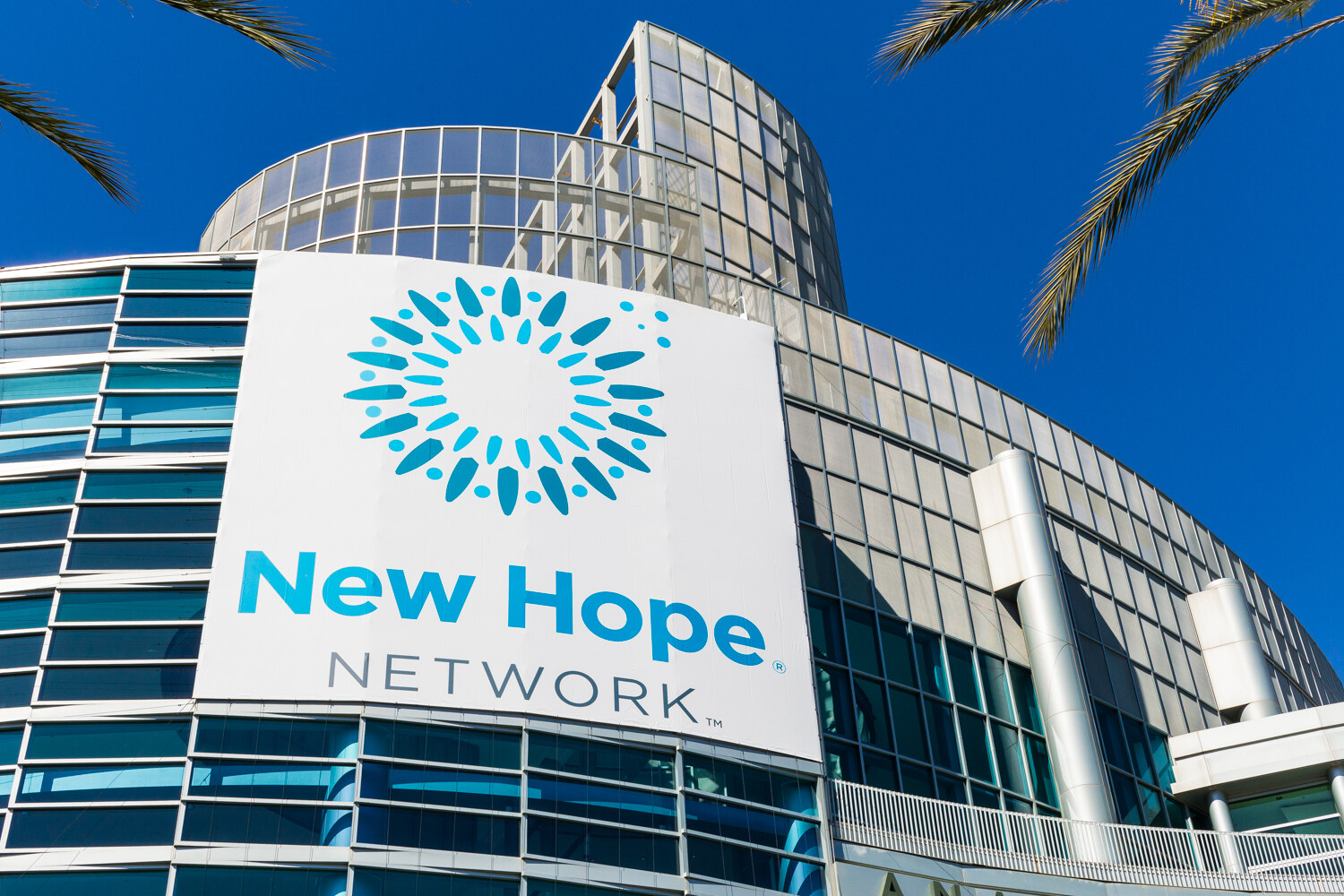 White banner with the blue starburst New Hope Network logo on a glass building, framed by palm fronds.