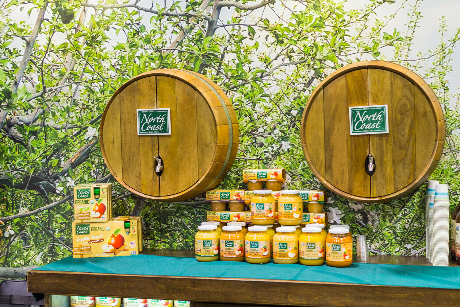 Table of North Coast organic applesauce with two wooden barrels carrying North Coast signs against an apple-tree mural.