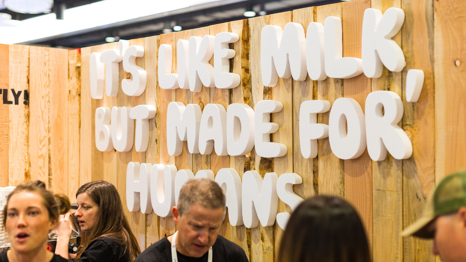 Wood wall with white foam letters reading It's like milk but made for humans, with several booth staff in the foreground.