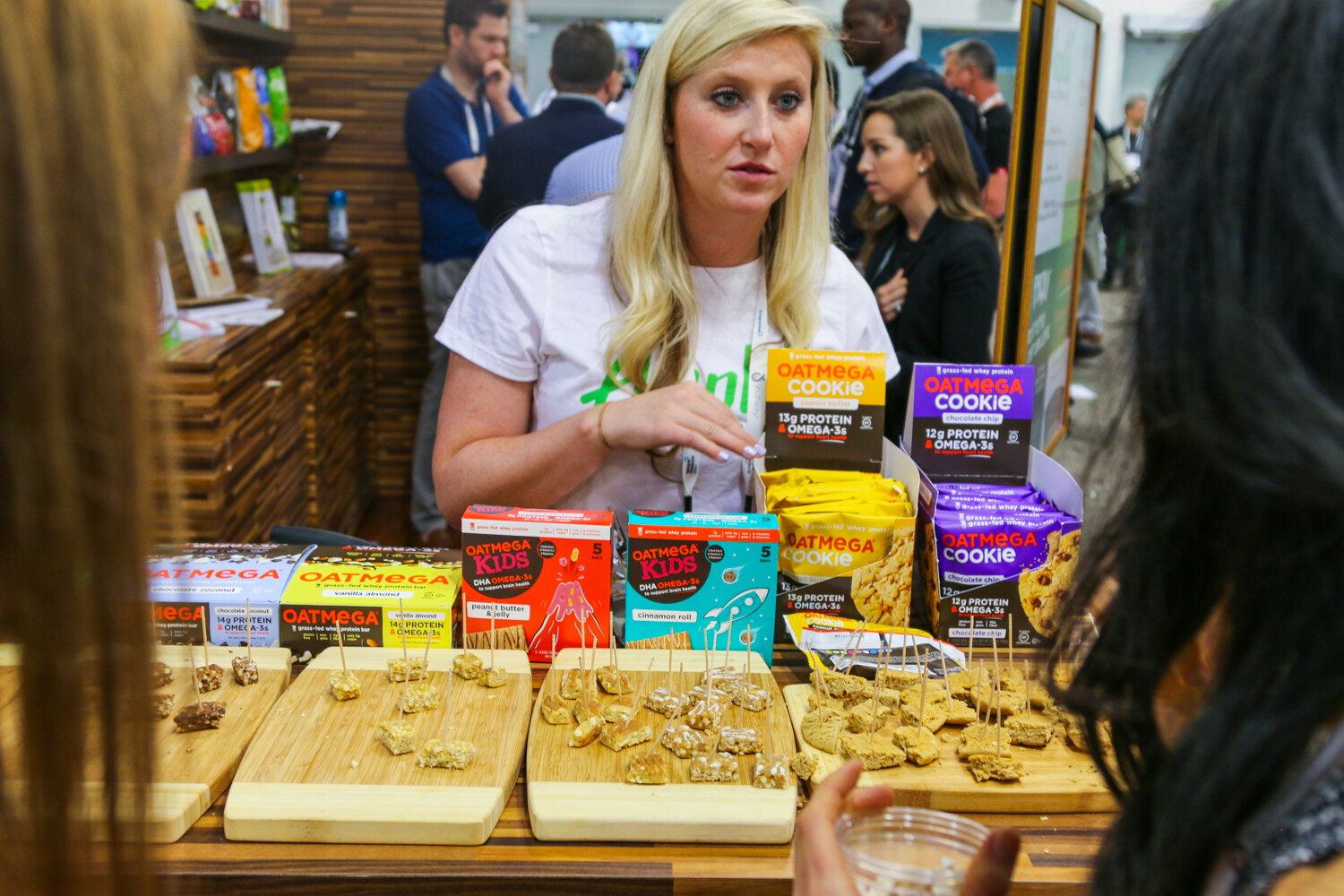 Blonde woman behind a wooden counter of Oatmega bars, Oatmega Kids and Oatmega Cookie boxes and skewered samples, with attendees in the foreground.