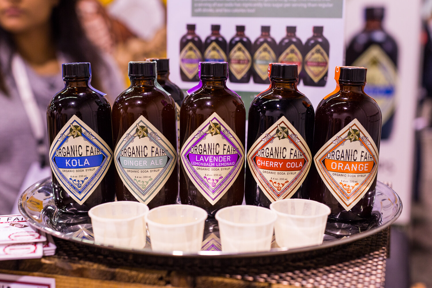 Brown bottles of Organic Fair soda syrup in kola, ginger ale, lavender lemonbalm lemonade, cherry cola and orange flavors with paper sample cups.