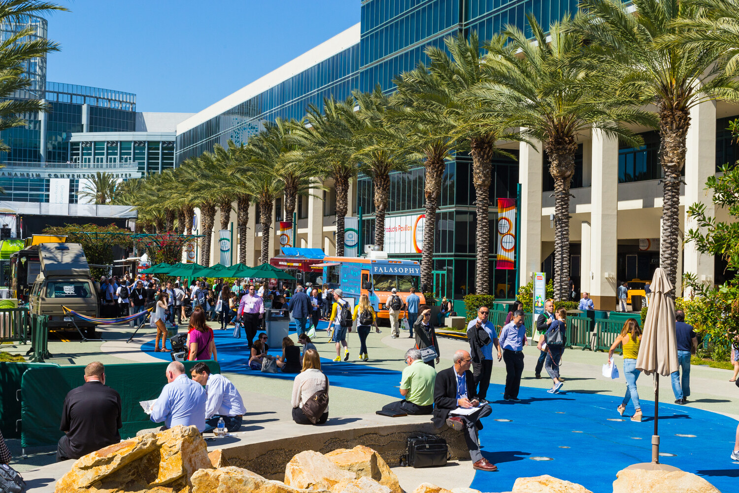 Wide view of a palm-lined plaza with attendees walking and sitting on a blue-painted pavement, with food trucks in the distance.