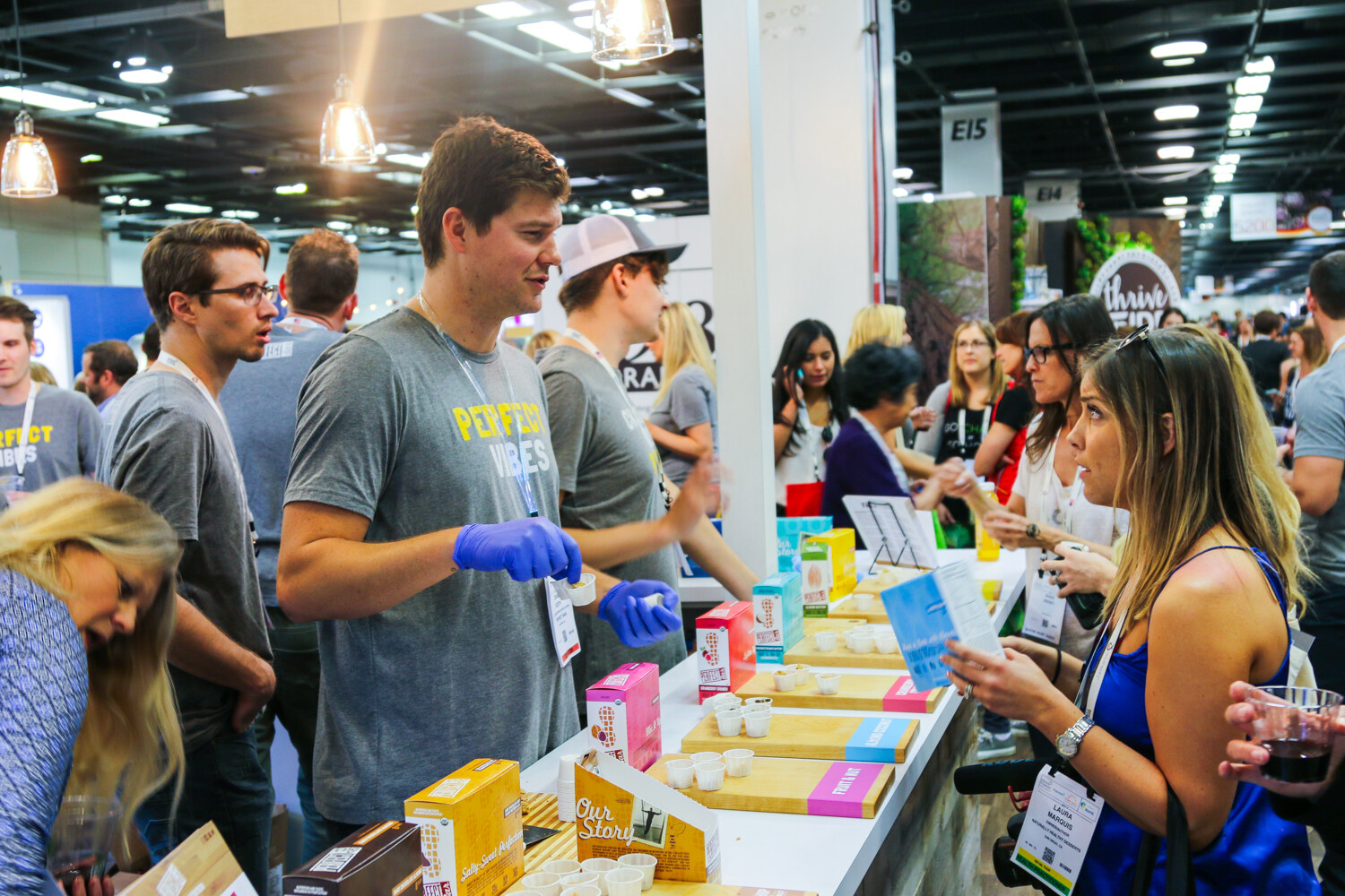 Staff in gray Perfect Bar shirts and a visitor in a blue dress at a sampling counter with boxes and small cups in a crowded aisle.